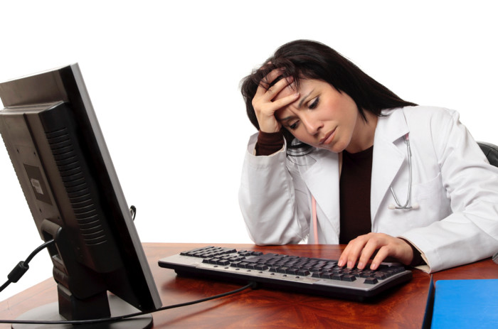 A physician in a white coat and stethoscope slumps at a desk, head resting wearily in one hand, staring at a bulky desktop monitor and keyboard.